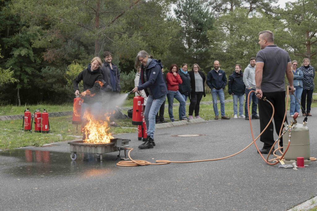 Teilnehmende einer Brandschutzhelfer Auffrischung löschen an einer Brandwanne im Freien mit Feuerlöschern, Trainer überwacht die Übung.