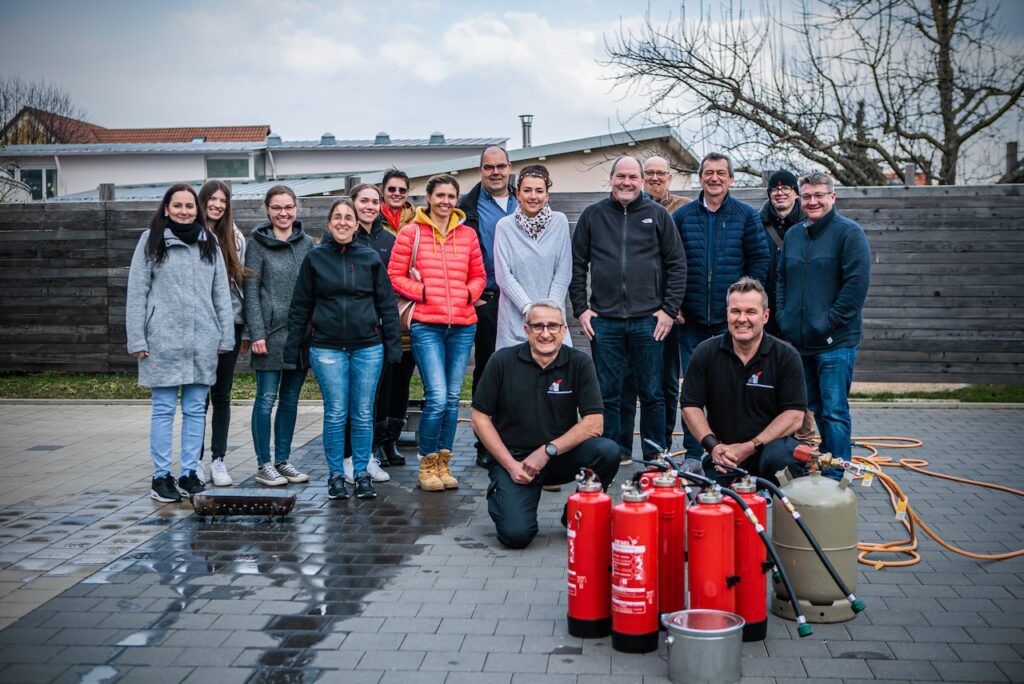 Teilnehmergruppe nach einer Brandschutzhelfer Auffrischung im Freien mit zwei Berufsfeuerwehrmännern und mehreren Feuerlöschern im Vordergrund.