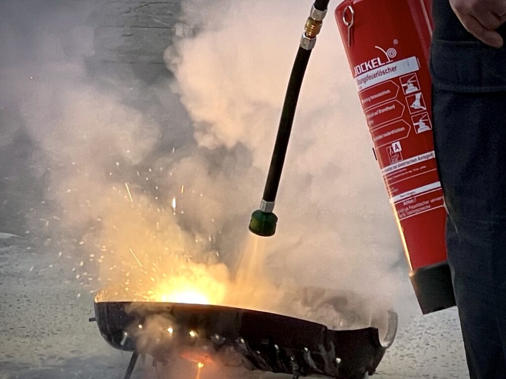 Echte Löschpraxis: Ein Feuerlöscher wird direkt auf eine offene Flamme abgegeben – Rückstoß, Hitze und reales Handling klar sichtbar.