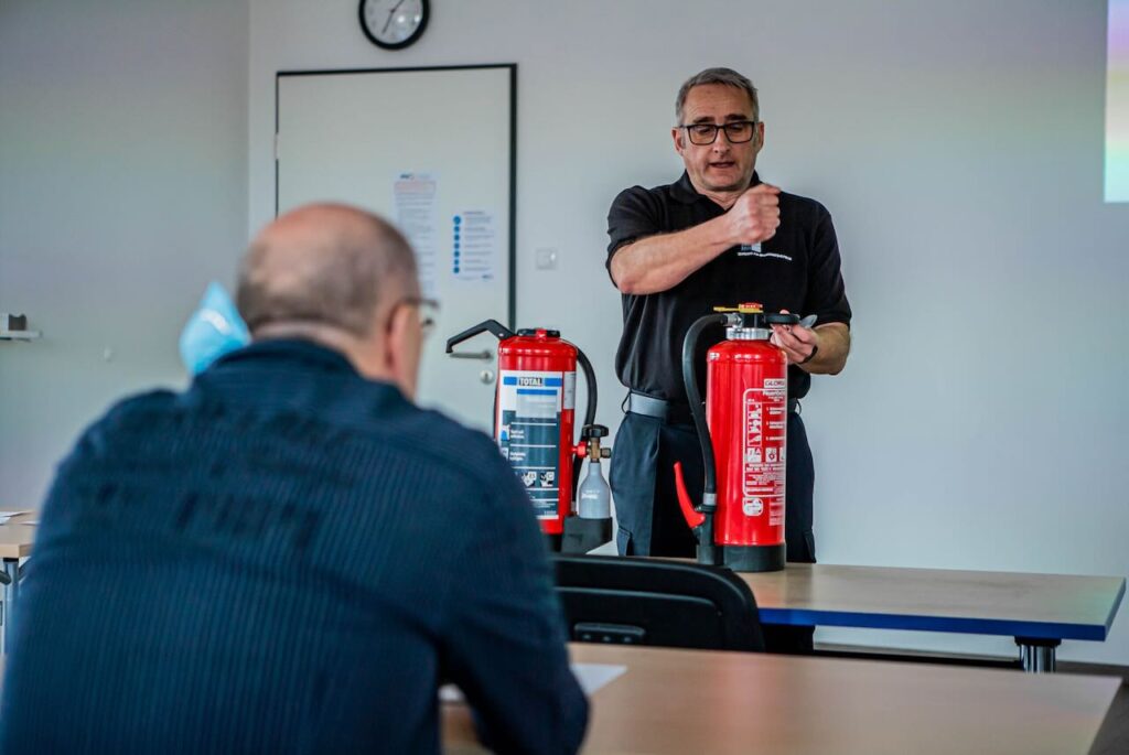 Berufsfeuerwehrmann erklärt im Seminarraum einer Gruppe Teilnehmender den richtigen Umgang mit Feuerlöschern – echte Ausbildung statt VR oder Online-Training bei der Brandschutzhelfer Ausbildung in Baden-Württemberg