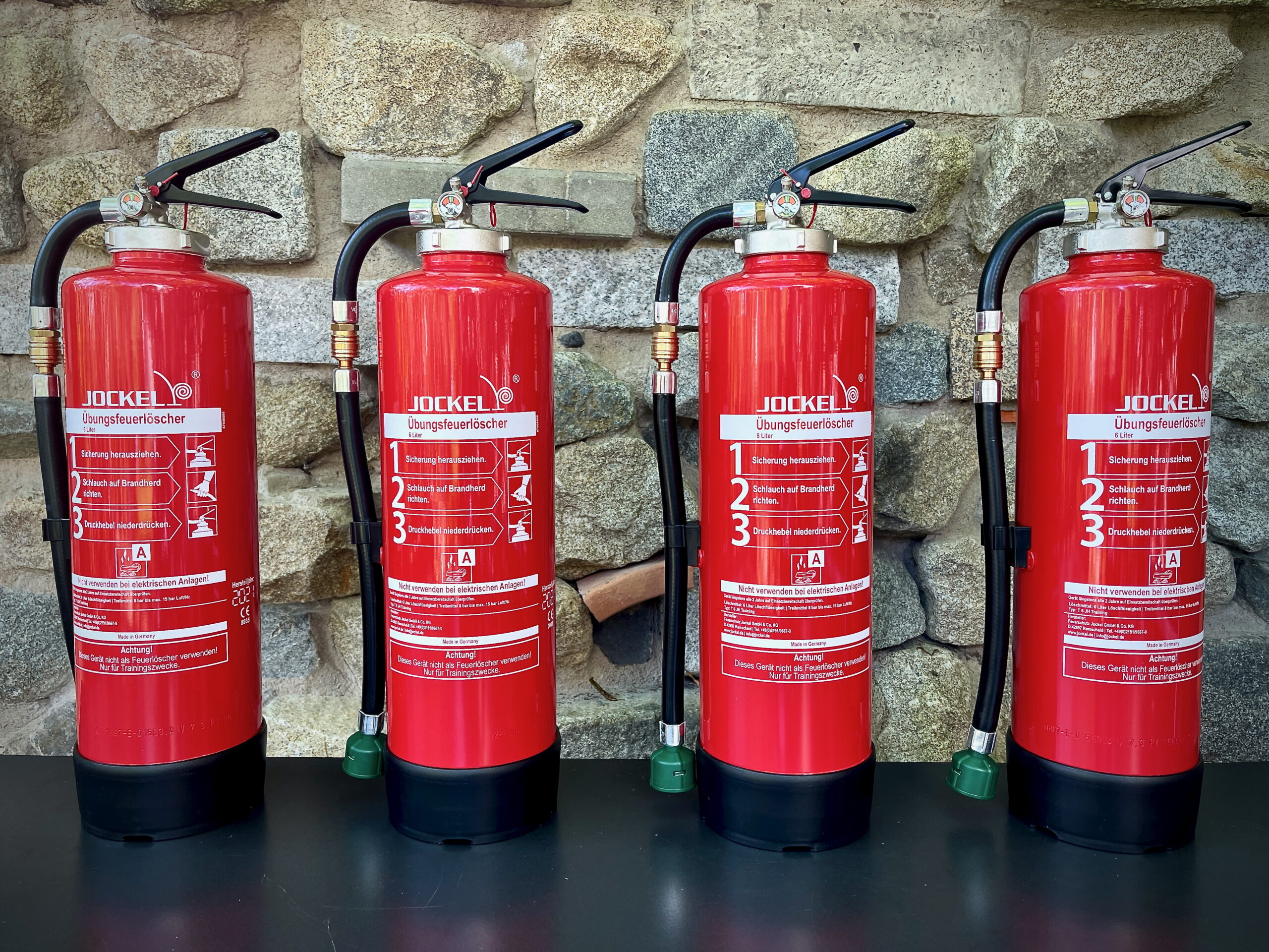 Four red fire extinguishers on a black table in front of a stone wall (photo, not AI-generated)