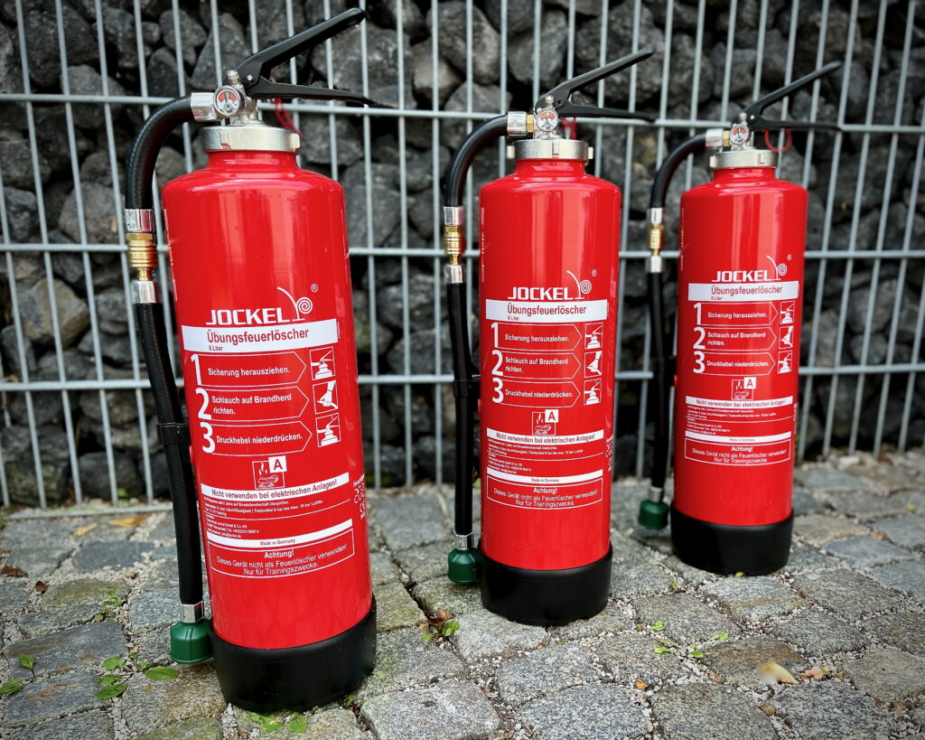 Three red training fire extinguishers by Jockel in an outdoor setting in front of a stone wall and metal fence (photo, not AI-generated)