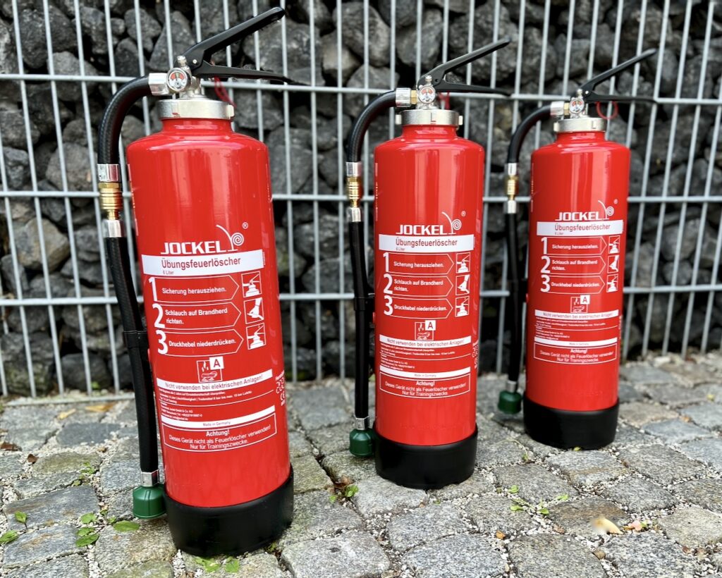 Drei rote Übungsfeuerlöscher der Marke Jockel in einer Außenaufnahme vor Steinwand