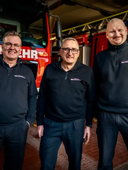 Von links nach rechts: Oberbrandmeister Thomas Till, Brandinspektor Heiko Schmidt und Gastdozent Brandinspektor Uwe Wahl vor einem Löschfahrzeug auf einer Feuerwache. Die drei Berufsfeuerwehrbeamten führen Brandschutzhelfer Auffrischungen für Unternehmen in Bayern und Baden-Württemberg durch.